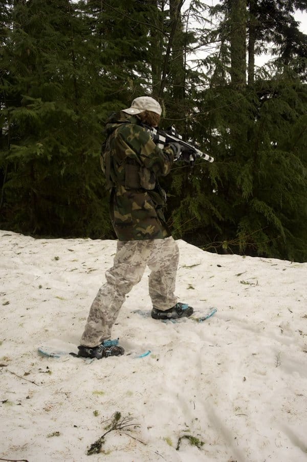 Individual in camouflage attire holding a rifle in a snowy forest setting.