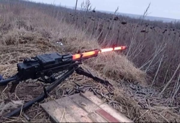A machine gun firing, emitting flames from its barrel, positioned on wooden planks in a barren, grassy field.