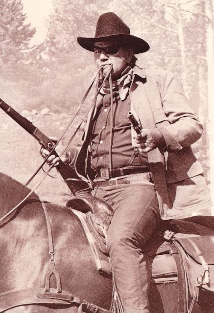 A man dressed in western attire riding a horse, holding a rifle and a pistol.