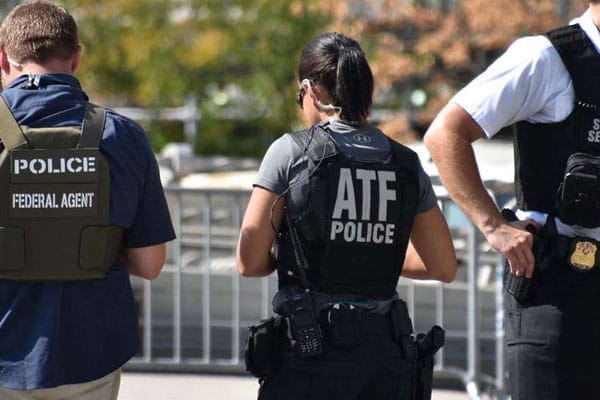 Three law enforcement agents, with two marked as "police" and one marked "atf police," seen from behind outdoors.
