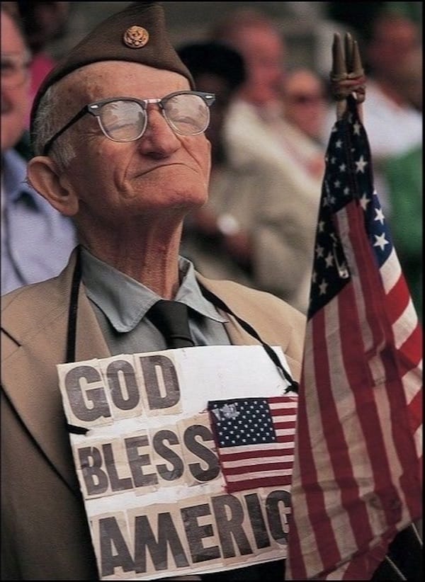 Elderly veteran wearing a beret, glasses, and a sign reading "god bless america," holding an american flag at a patriotic event.