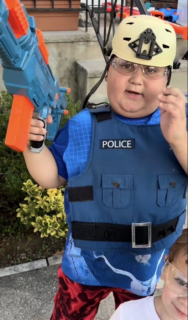 A child dressed in a play police vest and helmet holds a toy gun. Safety glasses rest on his forehead, and he is standing outside near greenery and concrete steps. Another child is partially visible.