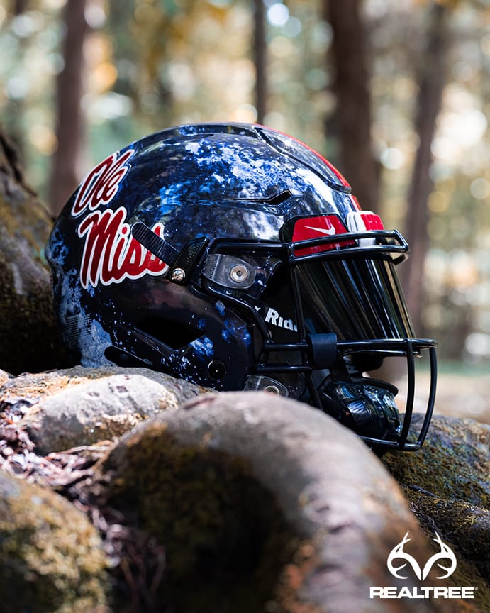 Football helmet with "Ole Miss" logo resting on a tree root. Forest background with sunlight filtering through leaves. Realtree logo in the bottom right corner.