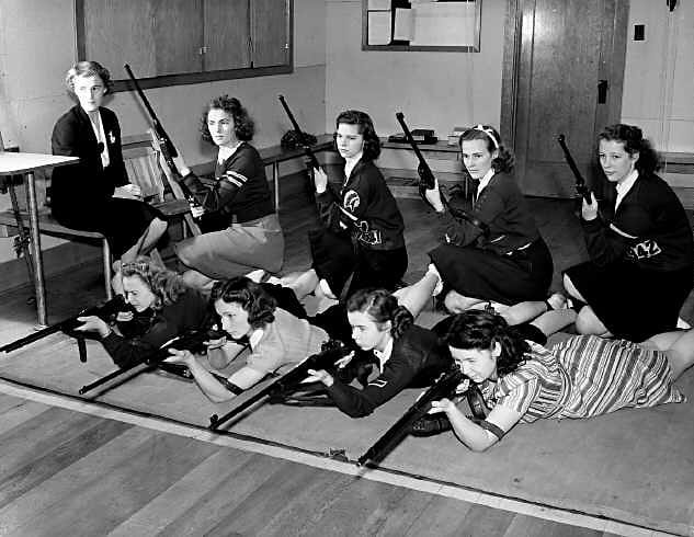 Nine women in a room holding rifles; four sitting and five lying on the floor, preparing for target practice.