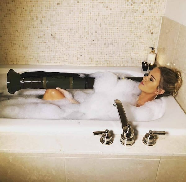 A woman is lying in a bathtub filled with bubbles, holding a large object that resembles a missile launcher. The bathroom has a modern design.