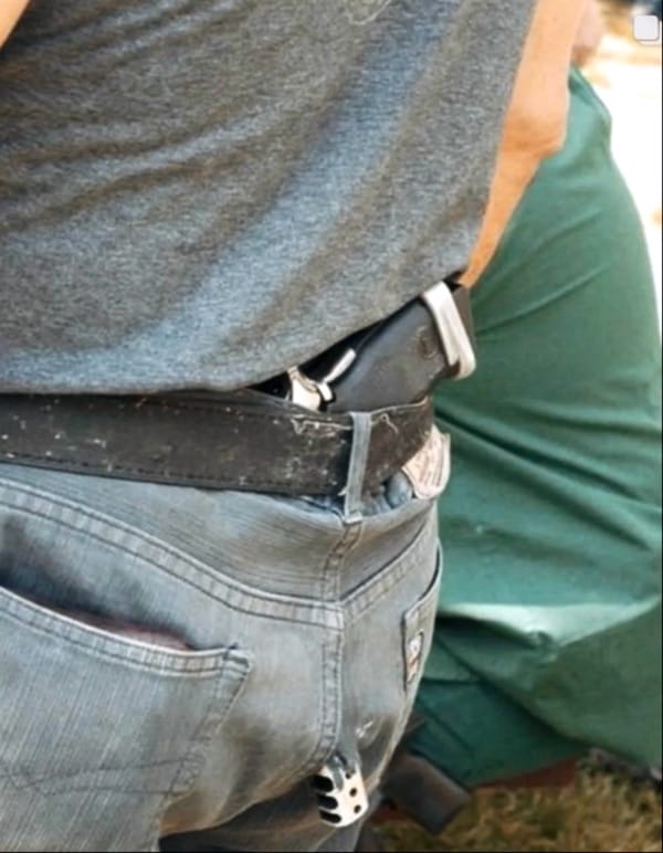 A person wearing a gray t-shirt and blue jeans with a black belt has a gun holstered at the waist.