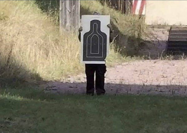 A person stands on grass holding a life-size shooting target in an outdoor setting.