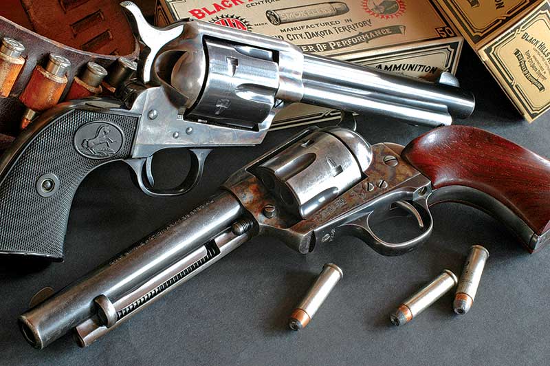 Two vintage revolvers are displayed on a surface surrounded by several bullets and ammunition boxes.